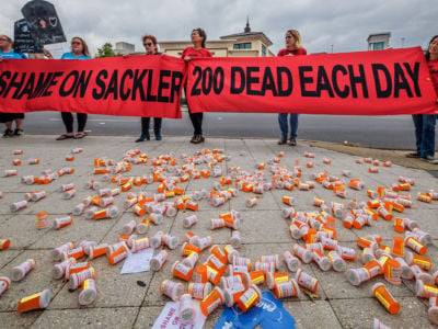 Activists hold a red banner reading "SHAME ON SACKLER; 200 DEAD EACH DAY" while standing in front of empty pill bottles scattered on the sidewalk