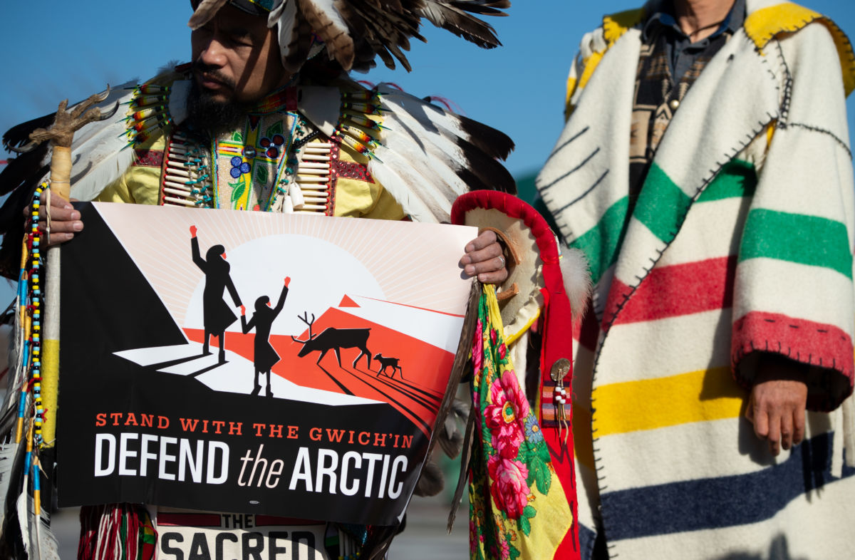 Indigenous activists hold a sign reading "STAND WITH THE GWICH'IN; DEFEND THE ARCTIC"