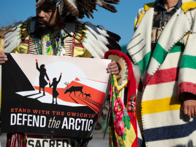 Indigenous activists hold a sign reading "STAND WITH THE GWICH'IN; DEFEND THE ARCTIC"