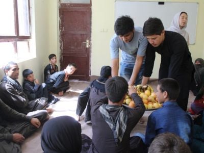 Two young men pass out fruit to people sitting in a room with them