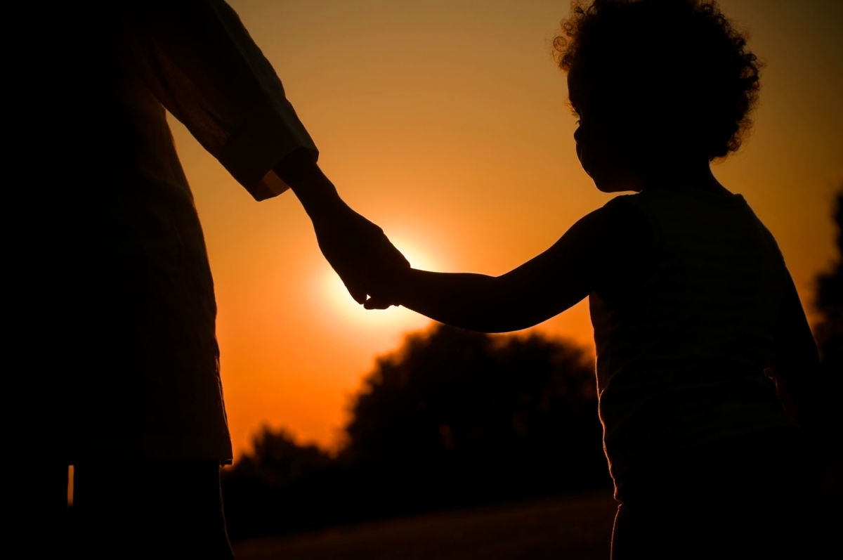 A Black child holds an adult's hands while silhouetted by the sunset