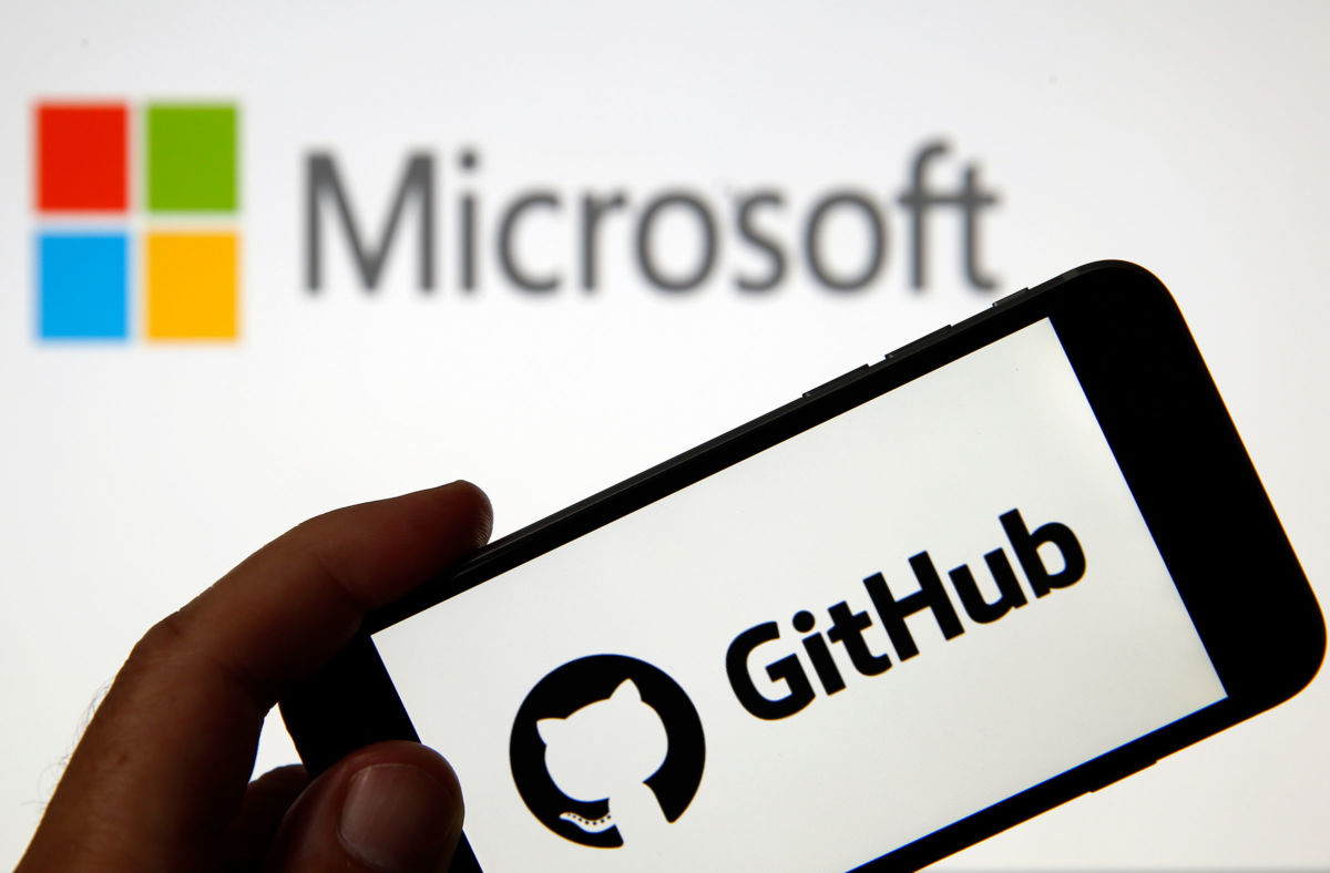 The GitHub logo is seen on the screen of an iPhone in front of a computer screen showing a Microsoft logo