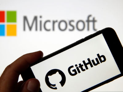 The GitHub logo is seen on the screen of an iPhone in front of a computer screen showing a Microsoft logo