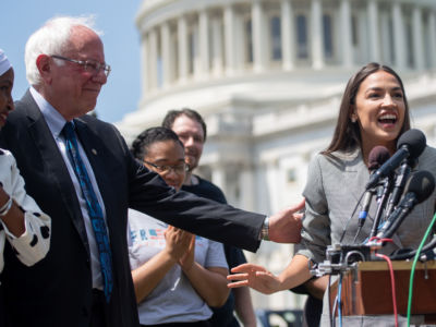 Bernie Sanders pats Alexandria Ocasio-Cortez on the back as she speaks at a podium in front of the US capitol