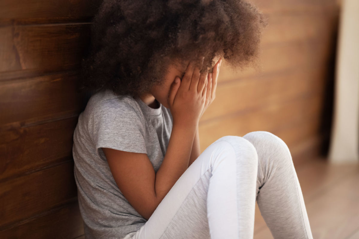 A young Black girl buries her face in her hands while sitting on the floor against a brick wall