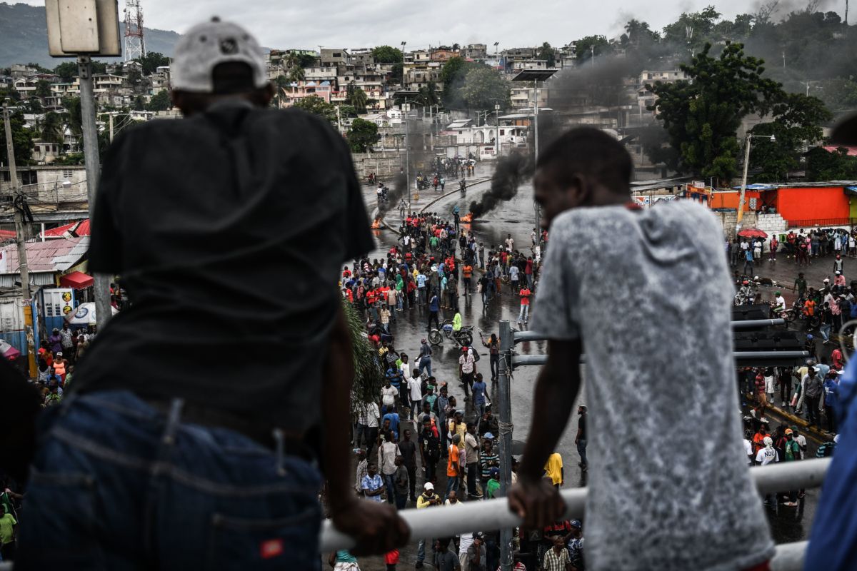 Two people stand on a balcony and look down on the street protest below them