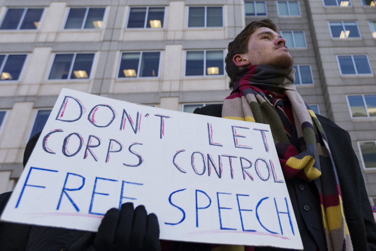 A man holds a sign reading "DON'T LET CORPS CONTROL FREE SPEECH"