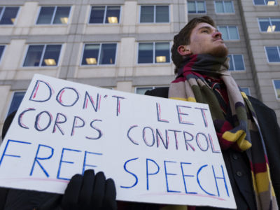 A man holds a sign reading "DON'T LET CORPS CONTROL FREE SPEECH"