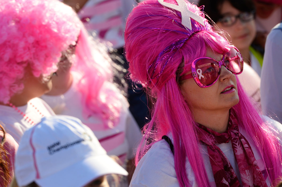 A woman in a pink wig with a pink ribbon painted on her sunglasses stands in a crowd with other runners