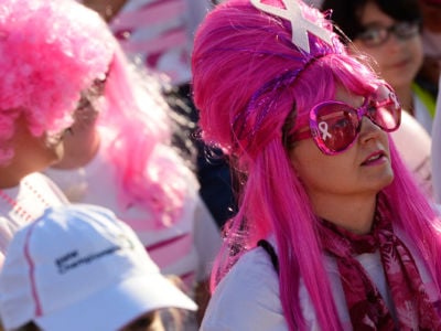 A woman in a pink wig with a pink ribbon painted on her sunglasses stands in a crowd with other runners