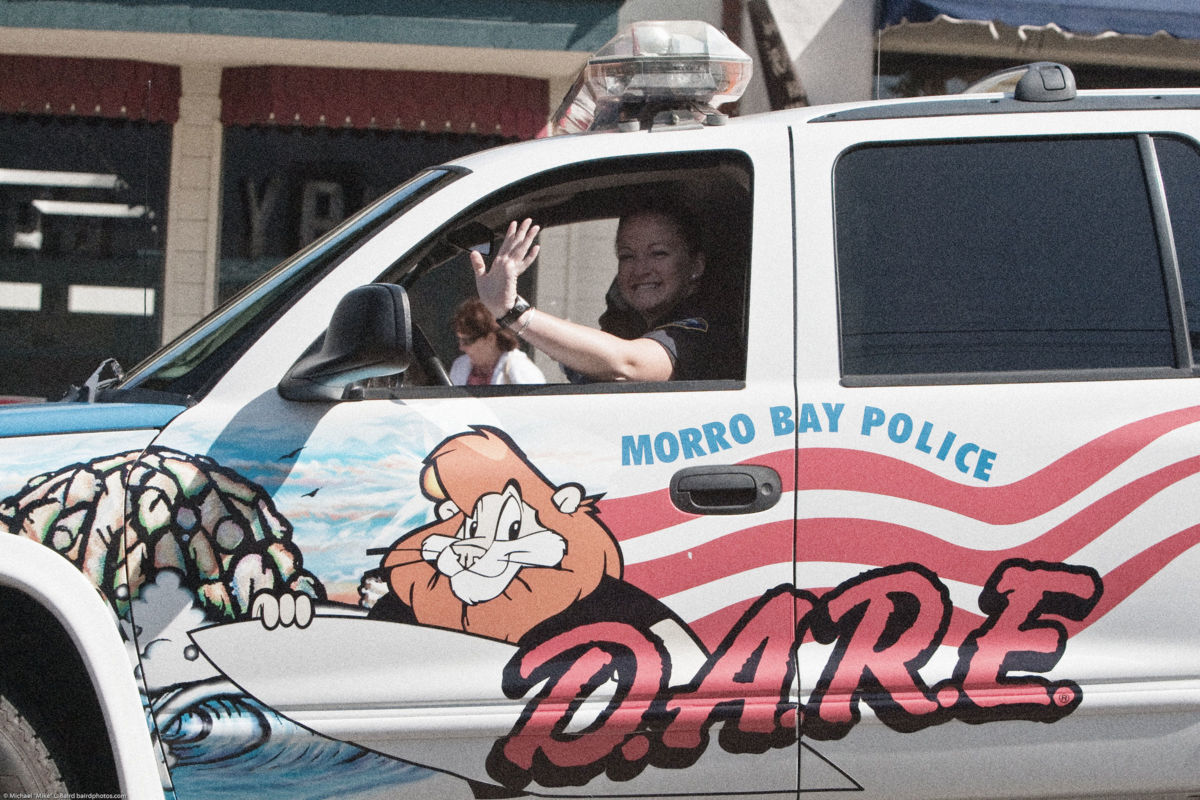 A woman waves from the drivers seat of a police SUV with DARE decorations, including a cartoon lion