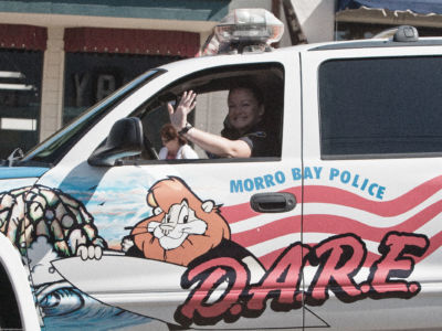 A woman waves from the drivers seat of a police SUV with DARE decorations, including a cartoon lion