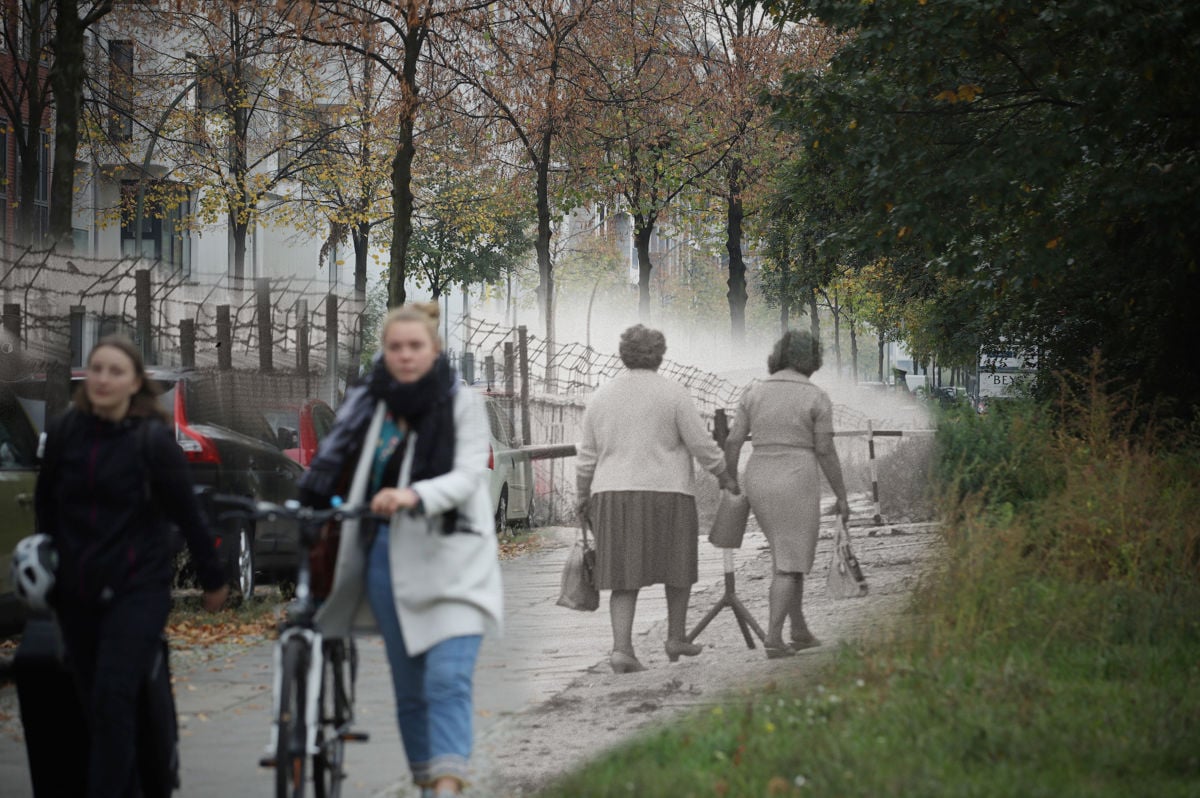 Two women walk in the foreground as another pair of women in black and white walk along the Berlin wall