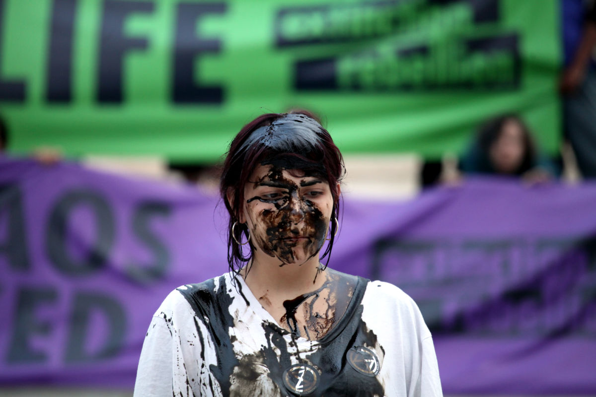 A woman covered in oil stands in front of a green Extinction Rebellion banner