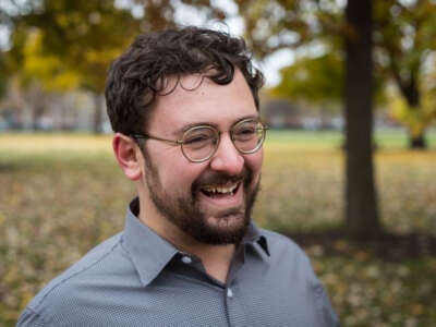 A photo of Ben Lorber a white man with round glasses and curly dark brown hair and beard. He is smiling