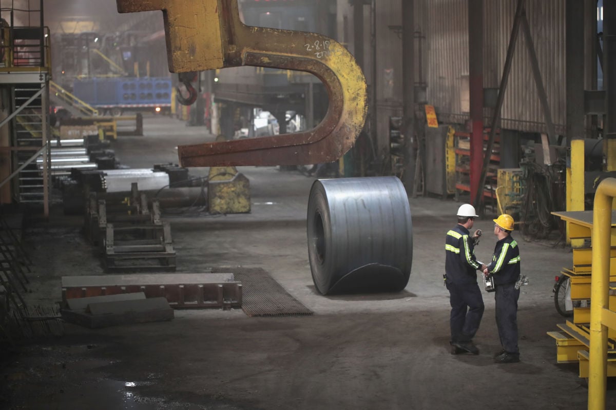 Two steel workers stand next to a large roll of steel in a factory