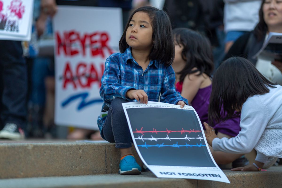 A young girl sits on the sidewalk during a protest while holding sign depicting red, white and blue lengths of barbed wire