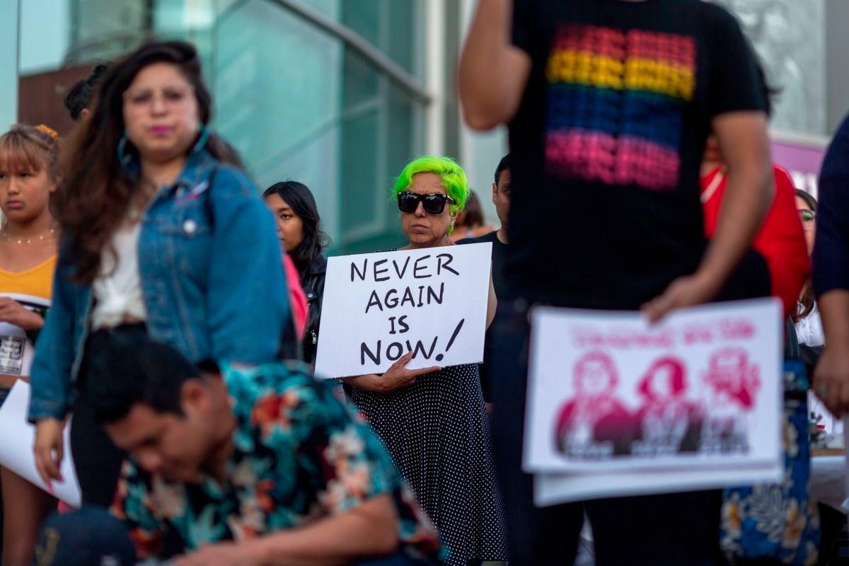 A woman with green hair and sunglasses holds a sign that reads "NEVER AGAIN IS NOW" during a protest