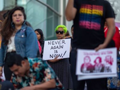 A woman with green hair and sunglasses holds a sign that reads "NEVER AGAIN IS NOW" during a protest