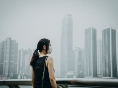 A young woman with a face mask on stands in front of an extremely smoggy city