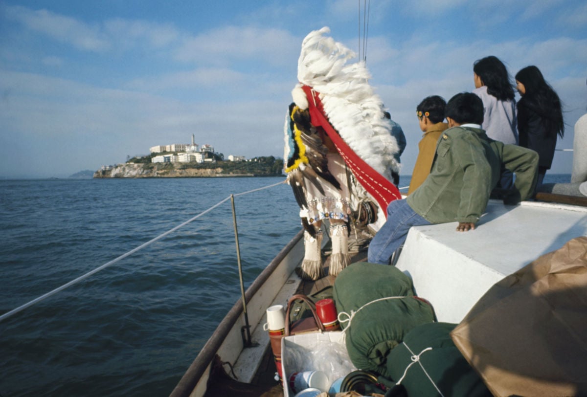 An elder in a war bonnet stands at the front of a motor boat headed to alcatraz