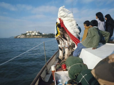 An elder in a war bonnet stands at the front of a motor boat headed to alcatraz