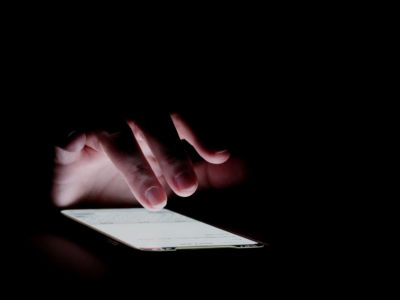 A hand taps on the lit screen of a smartphone in a dark room