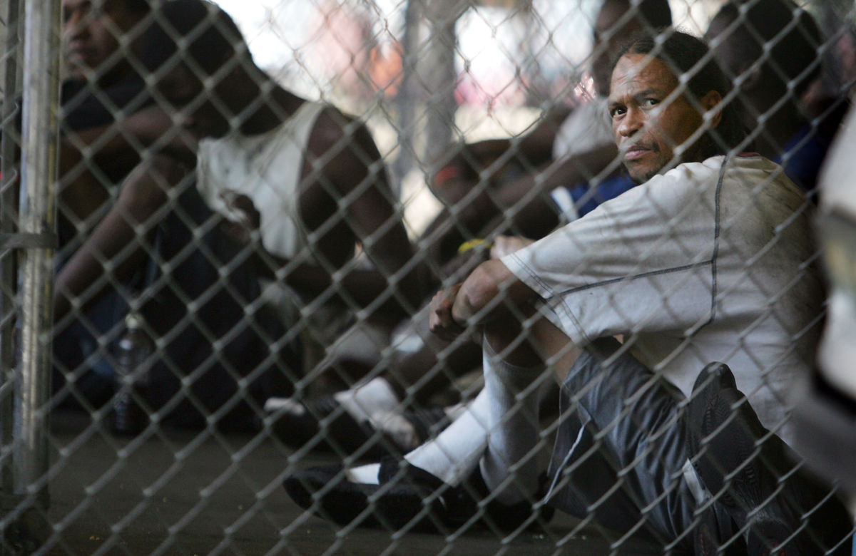 A man sits inside of a cage along with other prisoners