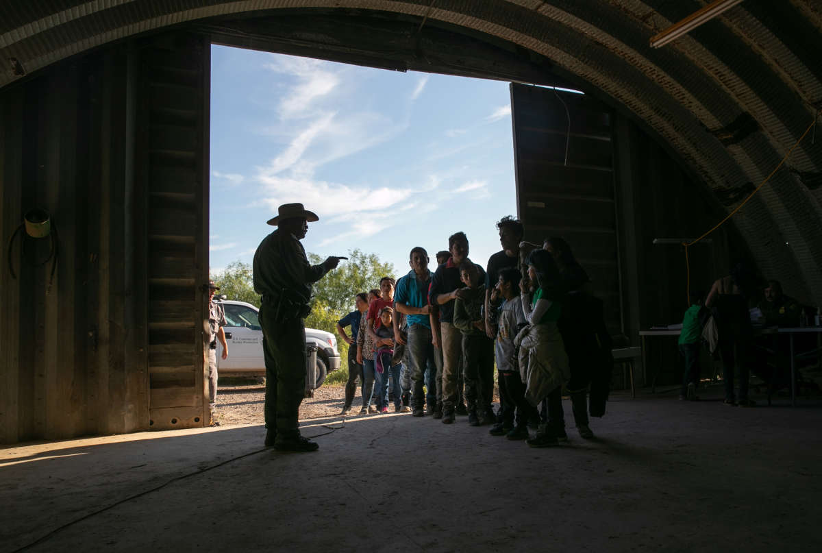 A seargant points his finger and speaks to migrants lined up in a domed shed