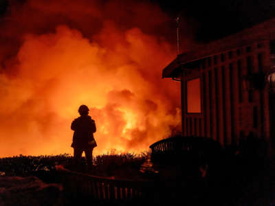 A person stands by a house and watchs a wildfire blaze