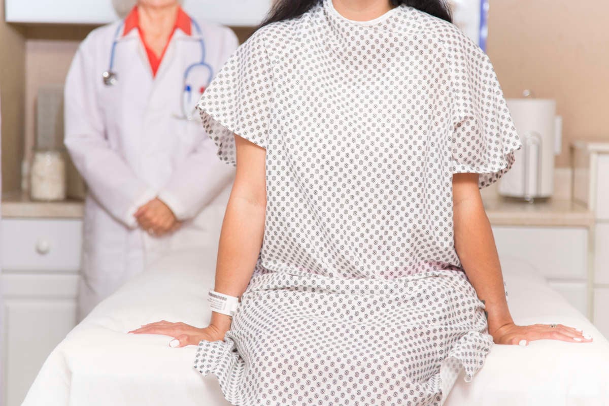 A woman in a hospital gown sits on a hospital bed as a doctor stands behind her