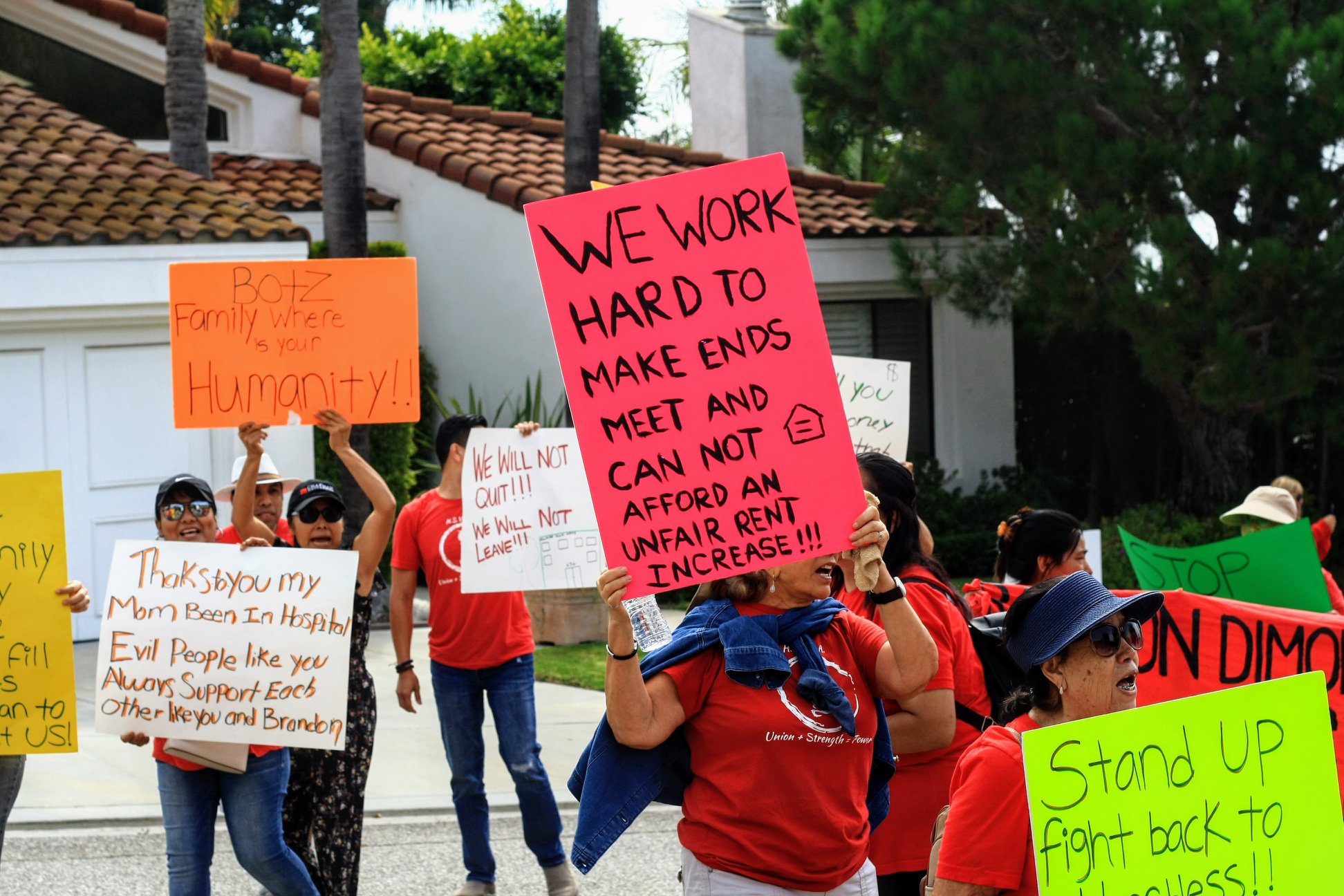 A protester holds a sign reading "WE WORK HARD TO MAKE ENDS MEET AND CAN NOT AFFORD A RENT INCREASE" while surrounded by others also displaying signs of their own