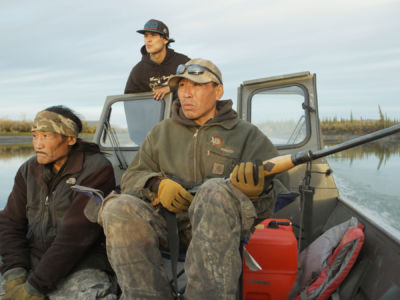 Three men, one of whom is carrying a rifle, sit in a moving boat