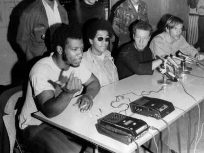 Activist Fred Hampton speaks into a microphone while seated at a table with others