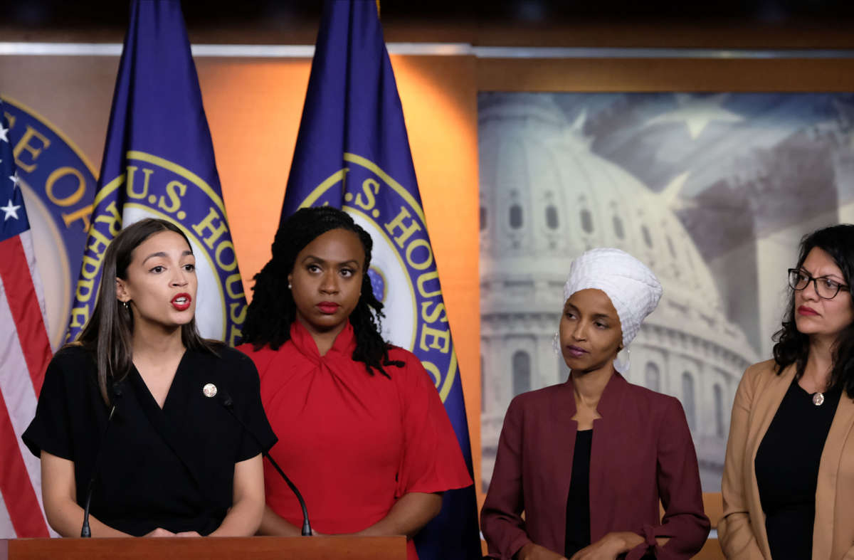 Alexandria Ocasio-Cortez speaks into a microphone while Ilhan Omar, Rashida Tlaib and Ayanna Pressley watch