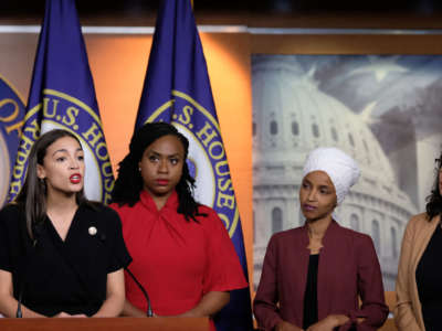 Alexandria Ocasio-Cortez speaks into a microphone while Ilhan Omar, Rashida Tlaib and Ayanna Pressley watch