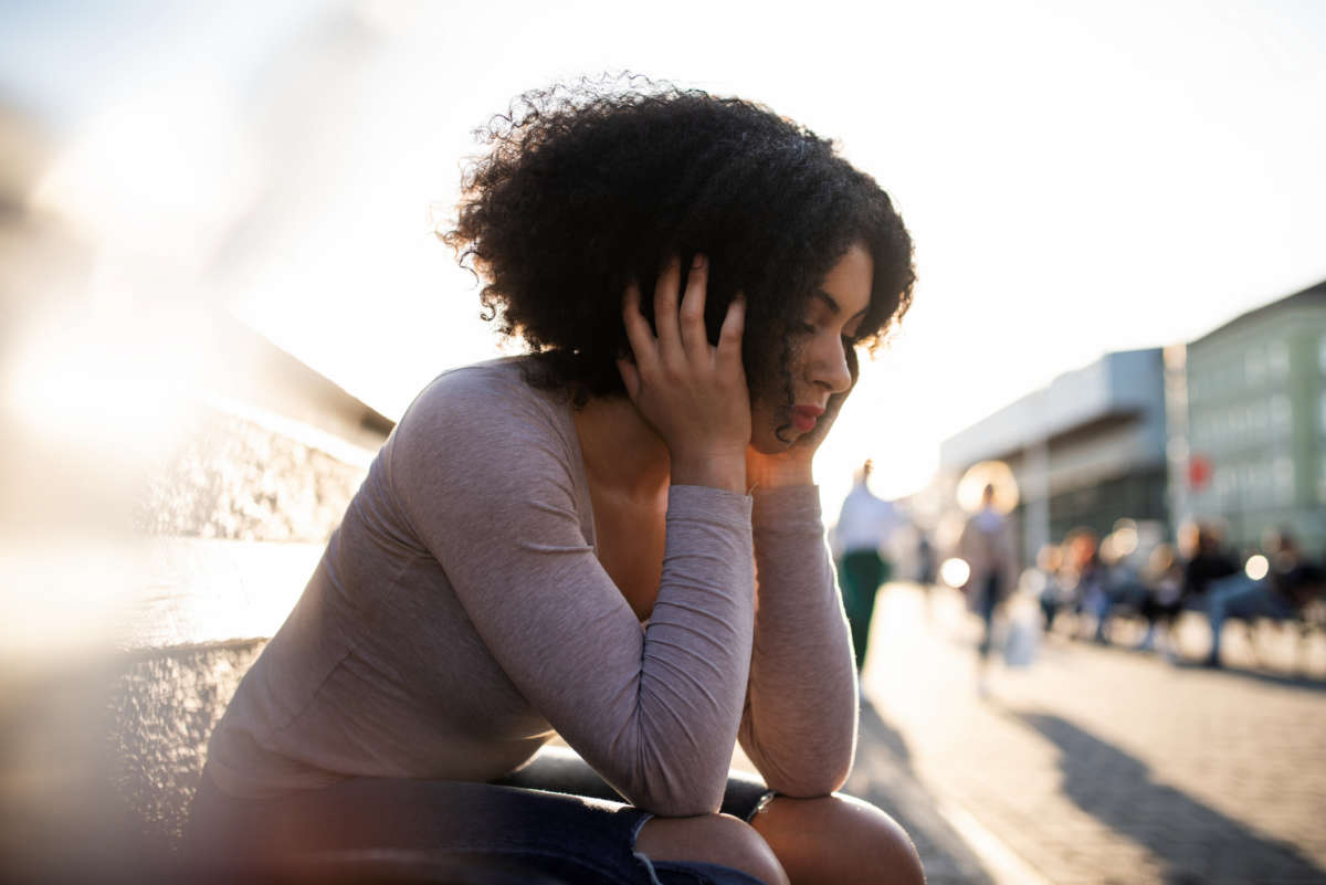 A woman sits on a bench outside holding her head in her hands