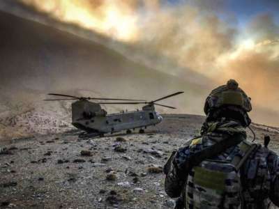 A soldier in fatigues looks at a helicopter on the dusty side of a mountain