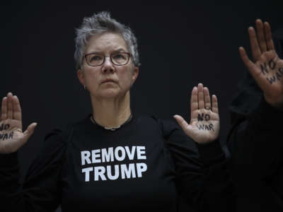 A woman wearing a shirt reading "REMOVE TRUMP" holds up her hands with "NO WAR" written on each one