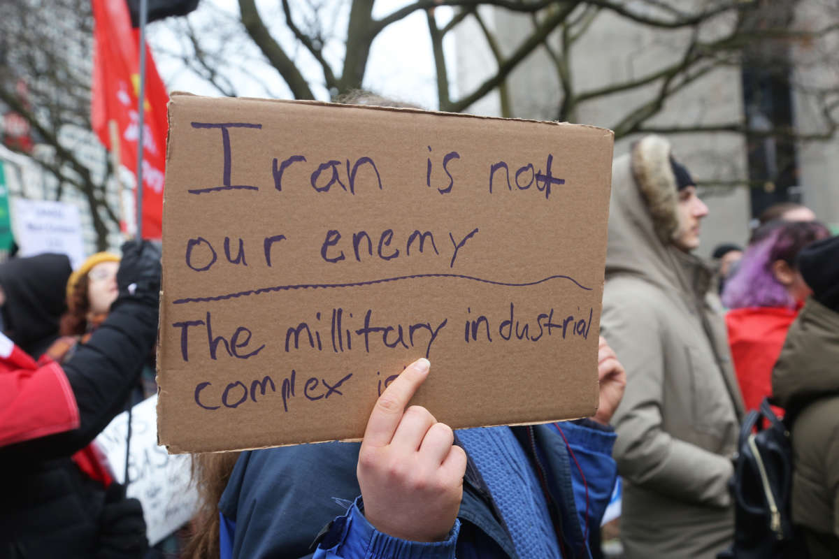 A protester holds a sign reading "IRAN IS NOT OUR ENEMY, THE MILITARY INDUSTRIAL COMPLEX IS"