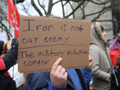 A protester holds a sign reading "IRAN IS NOT OUR ENEMY, THE MILITARY INDUSTRIAL COMPLEX IS"