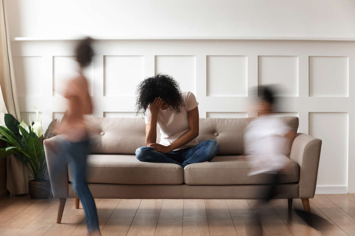 A black mother sits on a couch as her children run around her