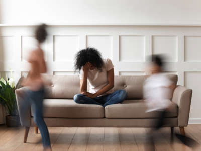 A black mother sits on a couch as her children run around her