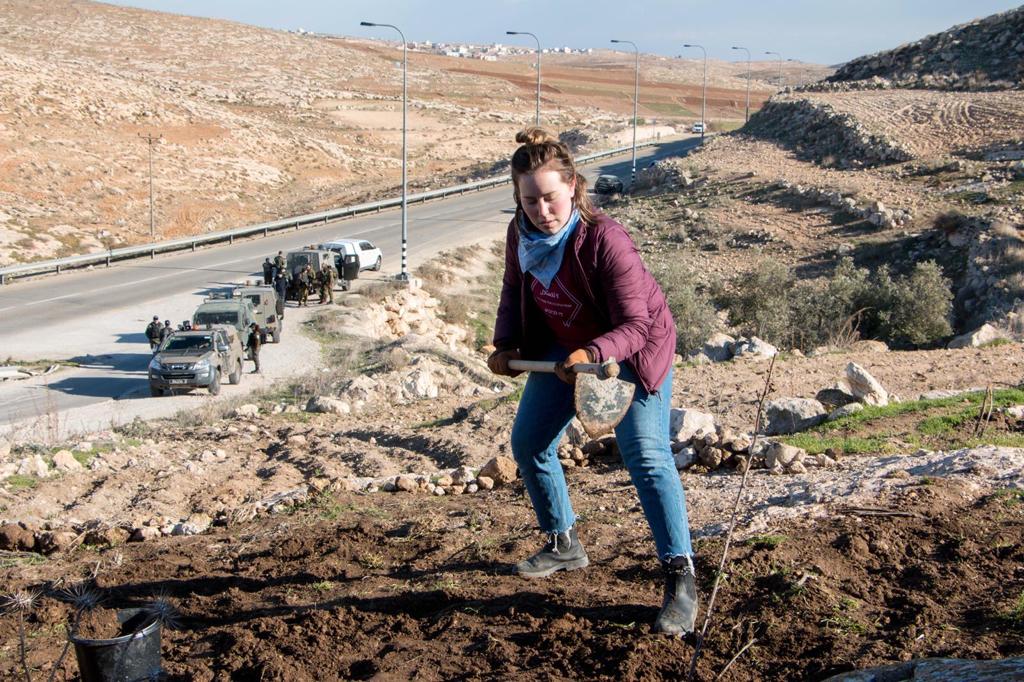 A woman with a farm tool works the ground as military vehicles line up in the distance behind her
