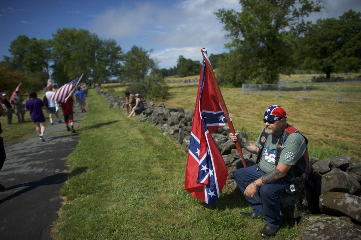 A man holding a confederate flag sits on a low wall