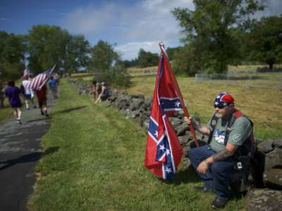 A man holding a confederate flag sits on a low wall