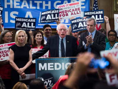 Bernie sanders speaks into a microphone surrounded by people holding "Medicare for All" posters