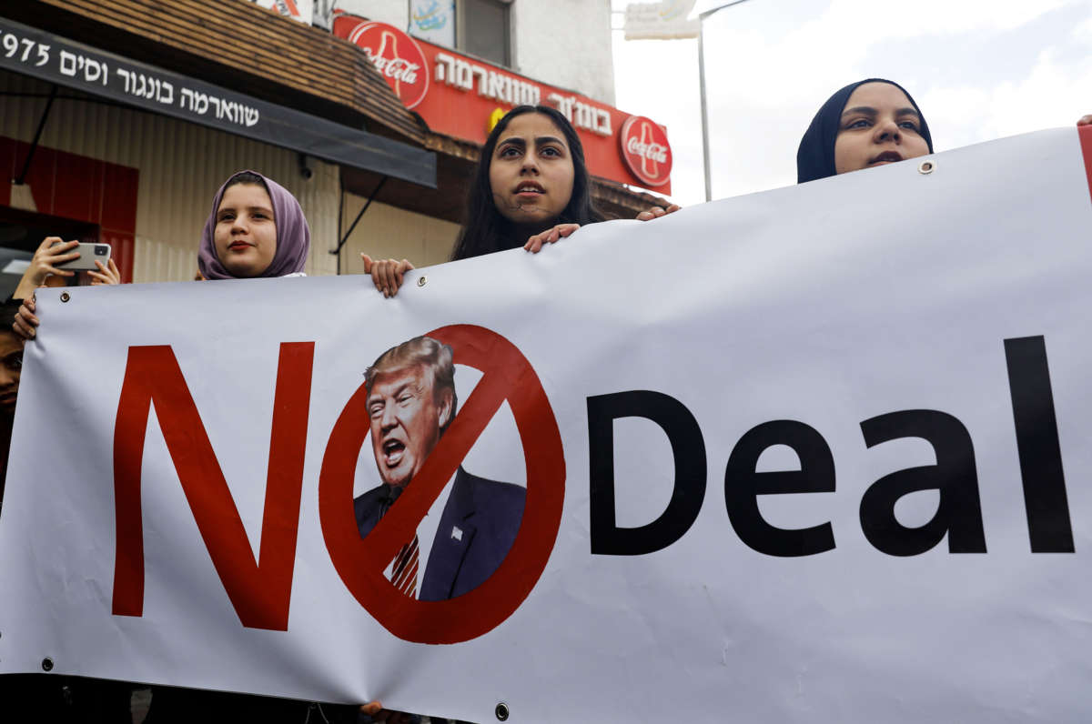 Three young women display a sign reading "NO DEAL," featuring Donald Trump with his face crossed out