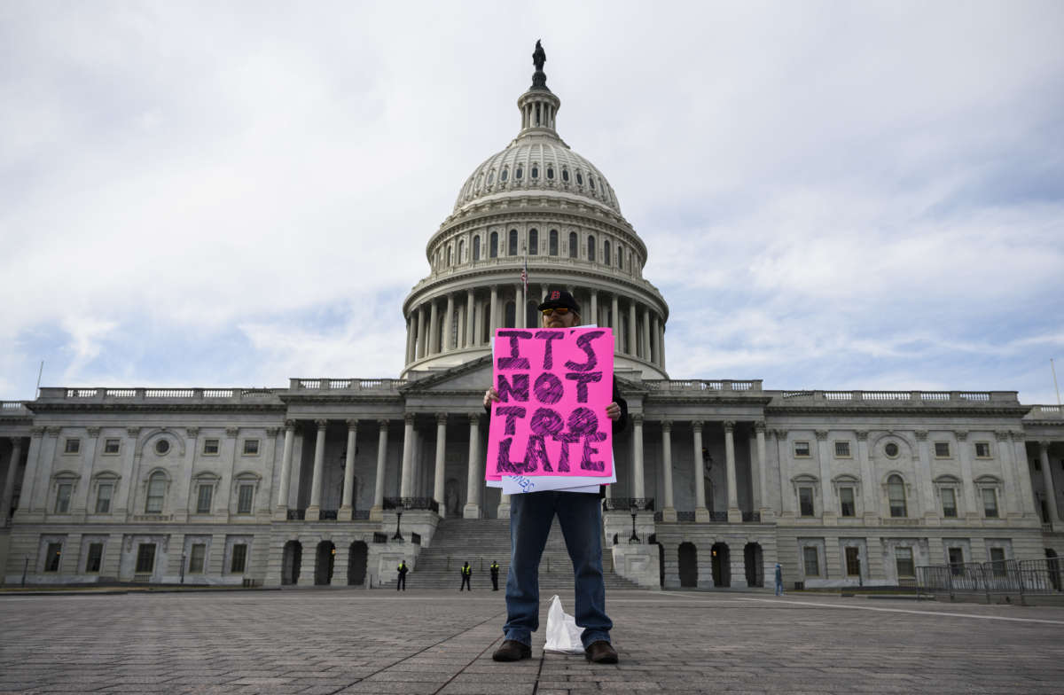 A lone protester stands on the steps of the capitol displaying a pink sign reading "IT'S NOT TOO LATE"