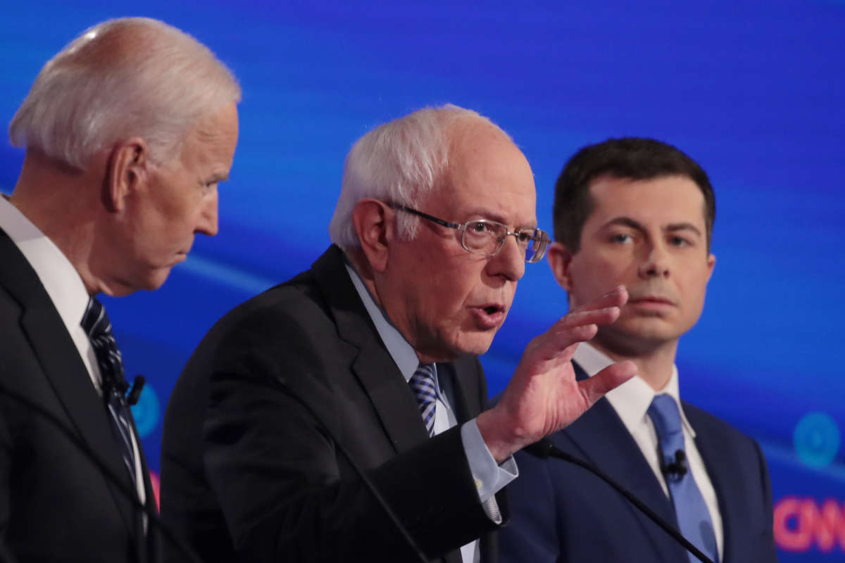 Bernie Sanders speaks during a debate while Joe Biden and Mayor Pete stand on either side of him at their respective podiums